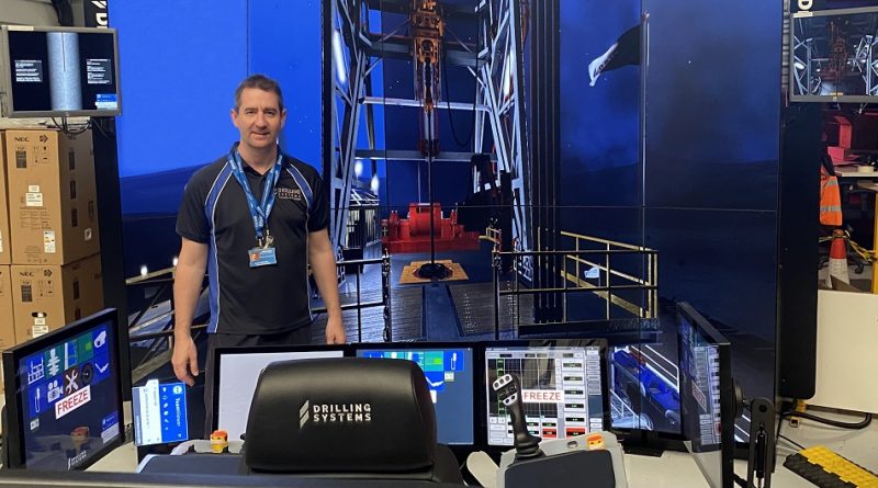 A man stands in front of a drilling simulation setup with multiple screens displaying drilling operations. He is wearing a blue shirt and a name badge. The area is well-lit with technical equipment visible.