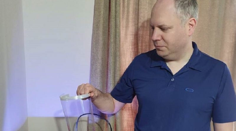 A man in a blue polo shirt looks at a transparent object with a handle against a beige curtain background.