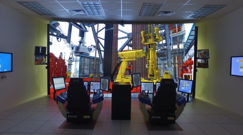 Two control stations with screens and joysticks face a large display showing an industrial machinery environment, likely simulating remote operations. Multiple monitors are mounted on the surrounding walls.