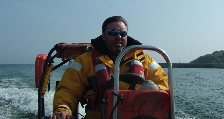 A man in a yellow jacket and blue sunglasses pilots a small boat on the water with a coastline visible in the background.