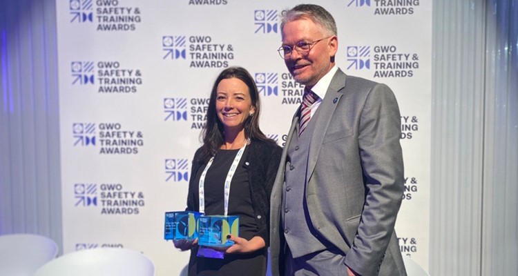 Two people stand smiling. The woman holds two awards, and a man stands beside her, both in business attire. Background has 