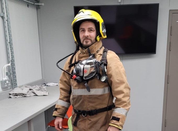 A person wearing firefighting gear, including a yellow helmet and breathing apparatus, stands in a room with a desk and a large screen on the wall.