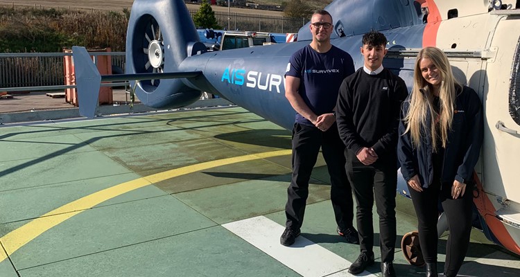 Three people standing on a helipad in front of a helicopter. The man on the left is wearing a navy blue shirt, the man in the middle is wearing a black outfit, and the woman on the right is in a navy blue jacket.