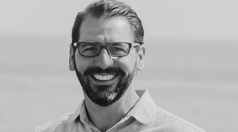 A man with short dark hair, a beard, glasses, and a light-colored shirt is smiling. The background shows a blurred expanse of water.