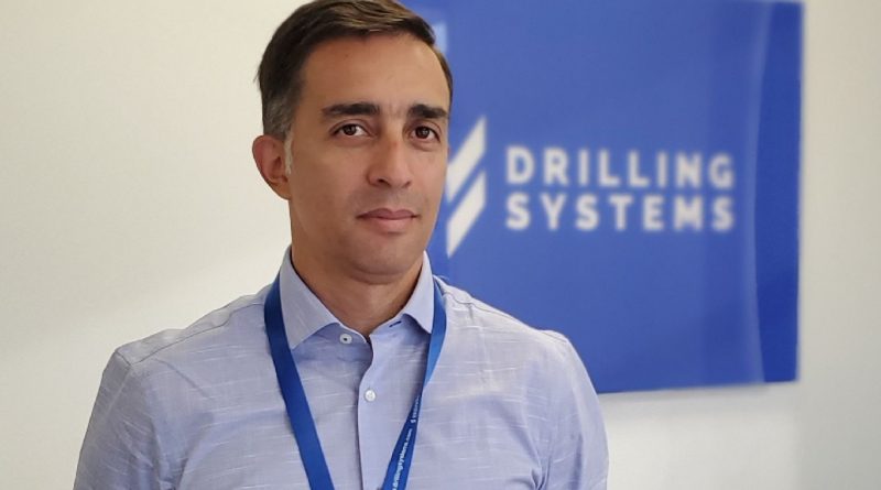 A man wearing a light blue shirt and blue lanyard stands in front of a blue sign that reads 