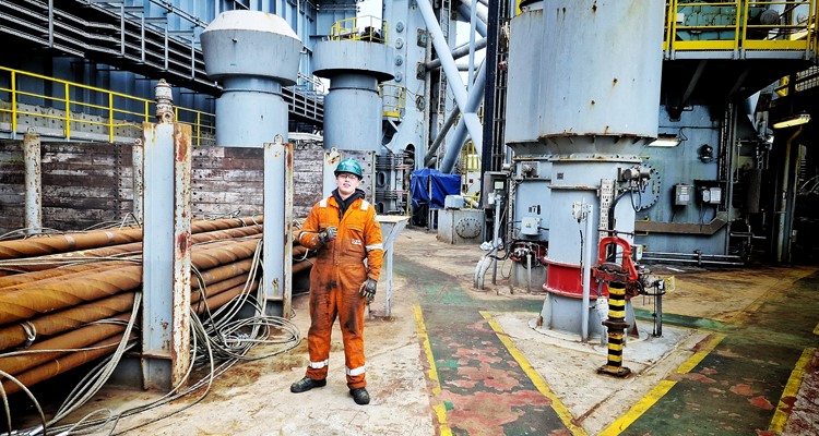 A worker in an orange jumpsuit stands on an industrial platform surrounded by large machinery and equipment.