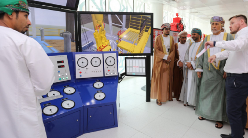 A group of men in traditional and Western attire gather around a control panel and display screens showing technical diagrams. One man in white points to the screens as he explains.