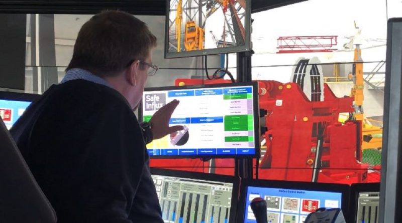 A man is sitting at a control panel with multiple monitors, gesturing towards one of them. A large piece of red industrial equipment is visible through a window.