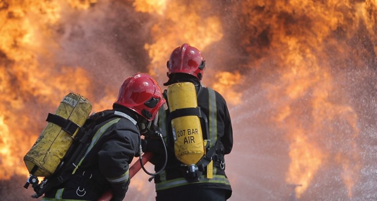 Two firefighters in protective gear and helmets combat intense flames, with water hoses directed at the fire.