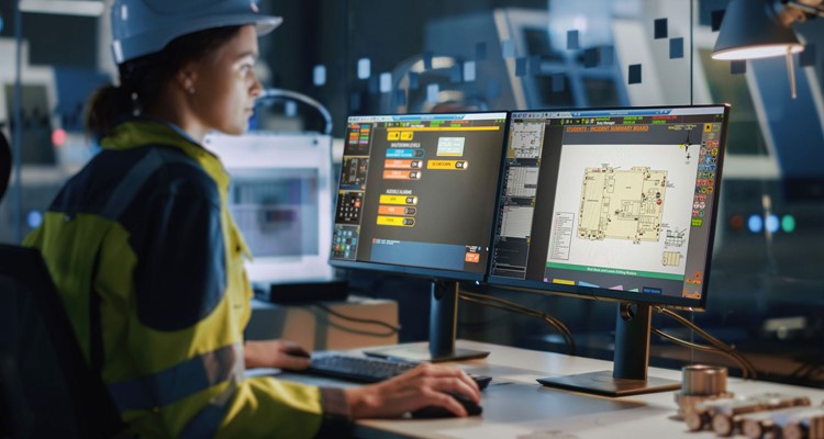 Person in a high-visibility jacket and hard hat works at a desk with two computer monitors displaying technical building plans and controls in an industrial setting.