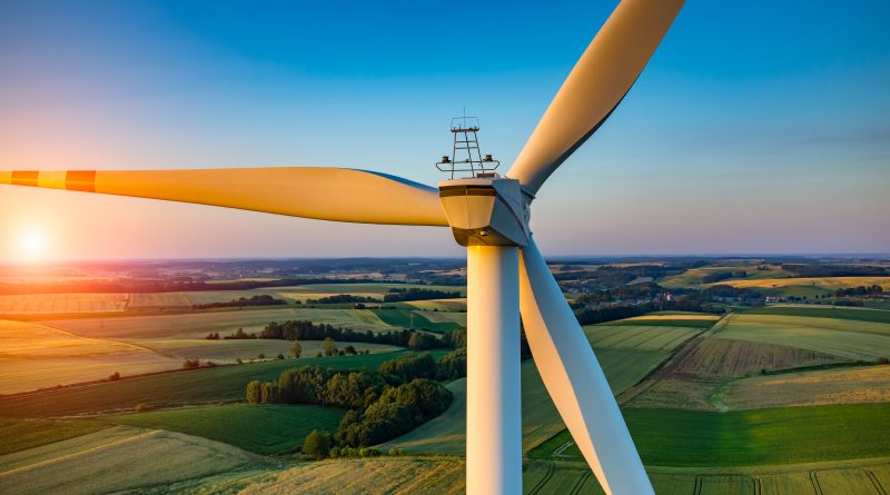 Close-up view of a wind turbine with a scenic countryside landscape and a setting sun in the background. Fields and hills are visible under a clear sky.