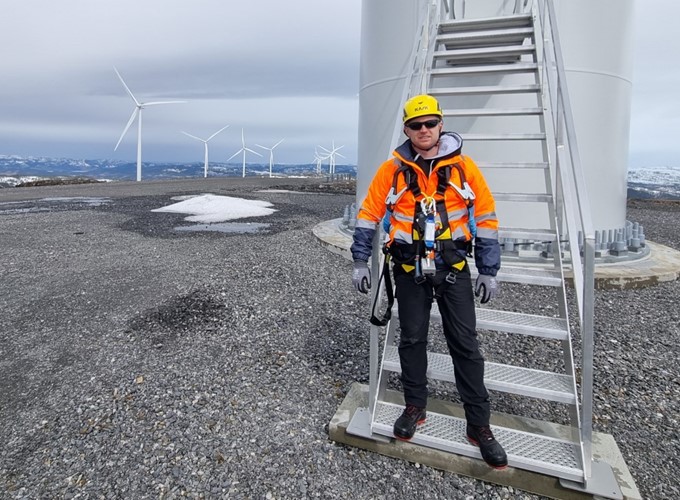 A person in safety gear stands at the base of a wind turbine, with multiple wind turbines visible in the background on a gravel-covered landscape.