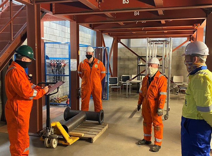 Four workers in safety gear have a discussion in an industrial setting, with materials and equipment visible in the background.