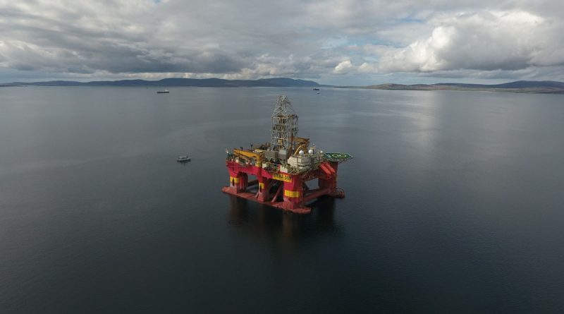 Offshore oil drilling rig floating in calm water under a cloudy sky with distant land and few boats visible.