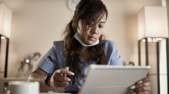 A person in a blue medical uniform, wearing a mask under their chin, is looking at a tablet and writing with a stylus.