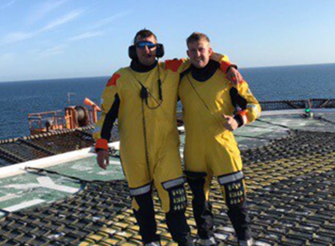 Two individuals in yellow protective suits stand side by side on a helipad offshore, with an expanse of the ocean and clear sky in the background.