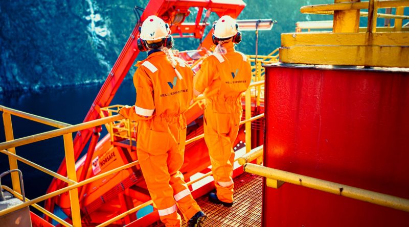 Two workers in orange safety uniforms and helmets standing on an offshore oil rig platform, facing away with equipment and safety railing visible around them.