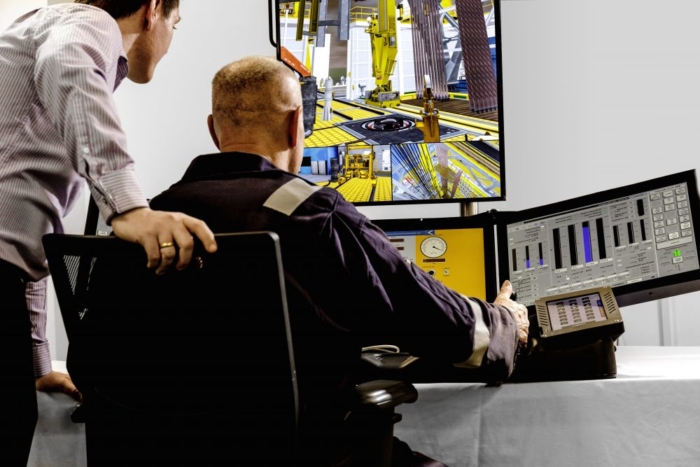 Two men in a control room monitor multiple screens displaying industrial machinery and data charts. One man sits while the other leans over, observing the screens.