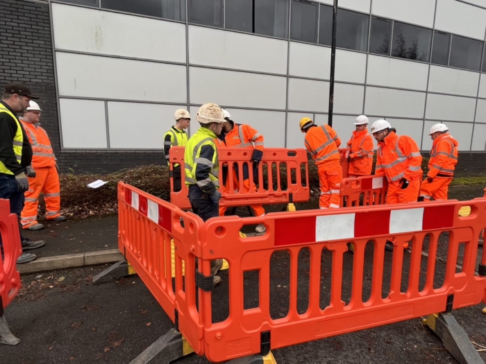 Construction workers in high-visibility clothing and hard hats gather around orange safety barriers on a street next to a building, demonstrating their expertise from comprehensive utilities and construction training.