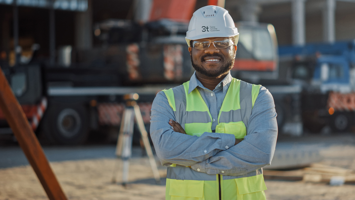 A smiling construction worker, donning a hard hat and safety vest, stands confidently with arms crossed at a construction site. Surrounded by equipment, he embodies the impact of rigorous training in ensuring a safe and efficient work environment.