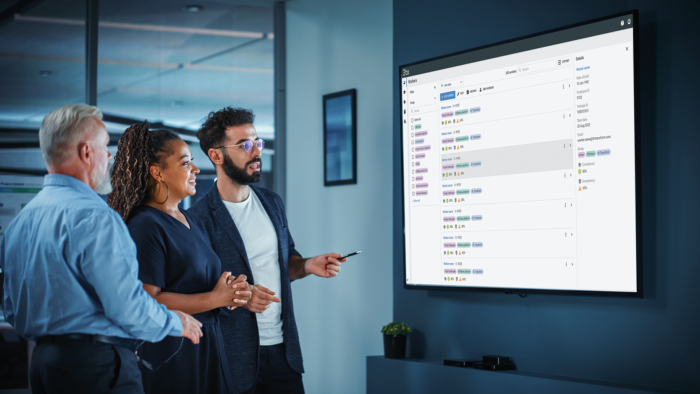 Three people stand in an office, looking at a large screen displaying a training schedule. One person is pointing at the screen.