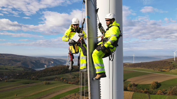 Two workers in safety gear, demonstrating their training, inspect a wind turbine while harnessed on ropes, with a scenic landscape of hills and more turbines in the background.