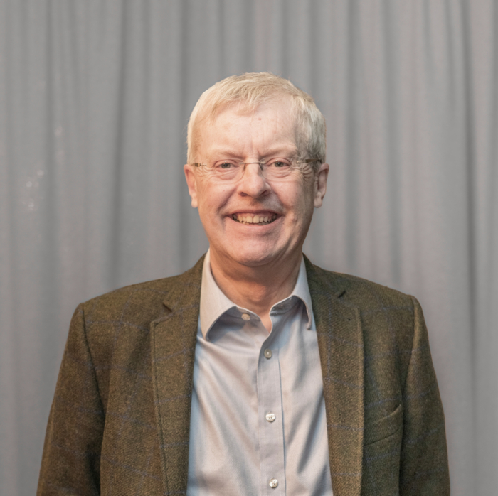 An older man with glasses and a gray jacket smiles confidently, possibly reflecting on years of training, in front of a gray curtain.