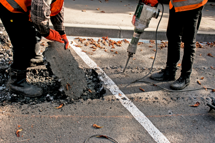 Two workers in safety gear, adept from rigorous training, break and remove a section of asphalt on a road using a jackhammer.