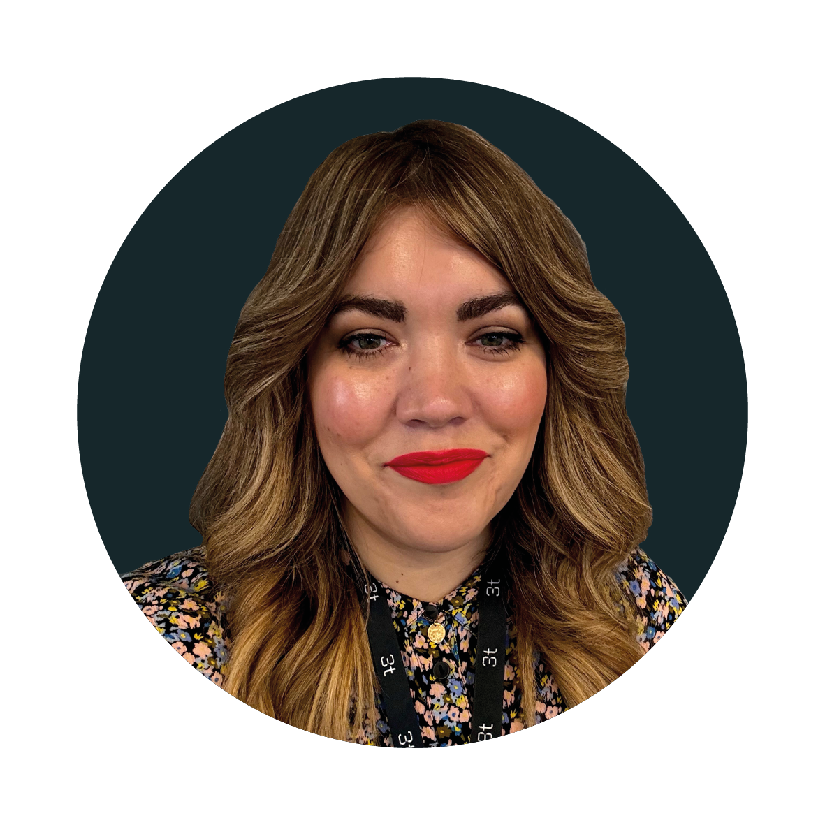 A woman with wavy brown hair and bright red lipstick, wearing a floral top and a black lanyard, smiles at the camera against a dark background.