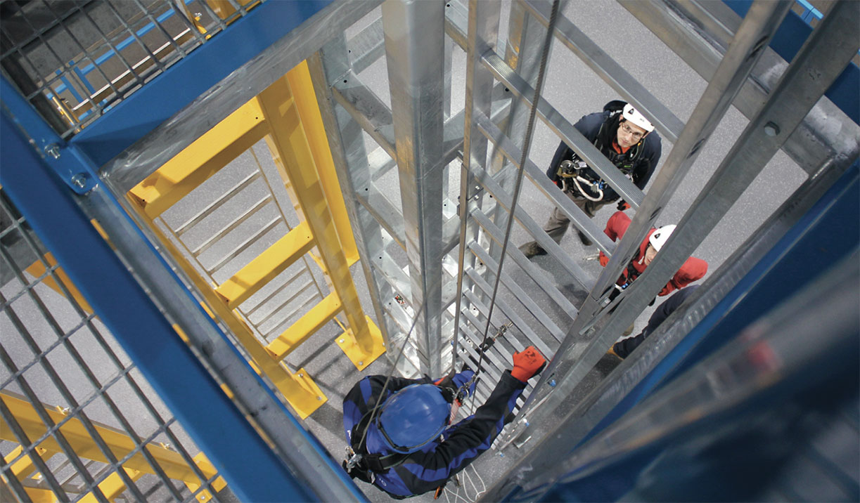 Workers equipped with safety gear are climbing a metallic industrial structure with ladders and platforms.