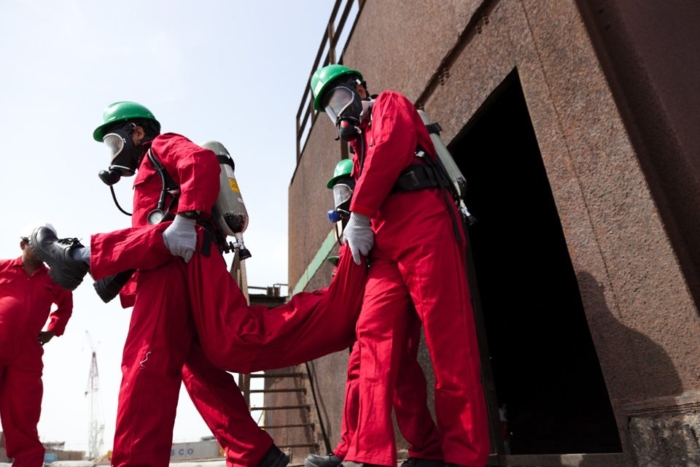 Emergency responders in red protective suits and green helmets carry an individual up a metal staircase.