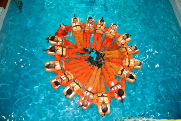 A group of people in orange suits and life jackets form a circle while floating in a pool, viewed from above.