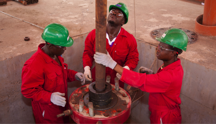 Three workers in red coveralls and green helmets are working with a large pipe in an industrial setting. Two are holding the pipe while one monitors, all wearing safety glasses and gloves.