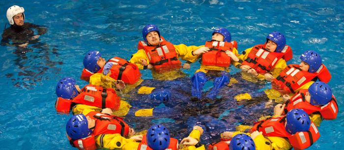 A group of people wearing life jackets and helmets form a circle while floating in a pool, resembling a strategic military formation. A person in a white helmet watches nearby.