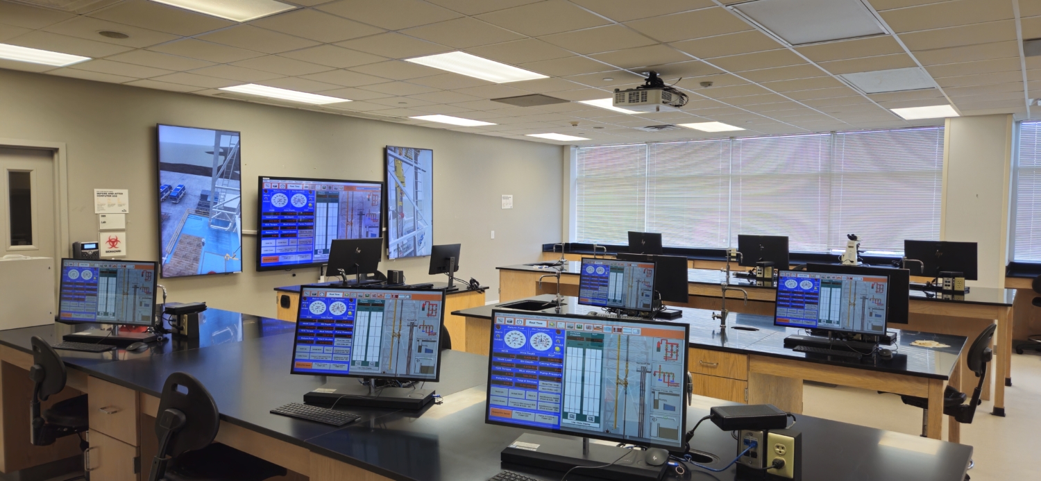 A modern control room with multiple workstations, each featuring monitors displaying control system interfaces. Large wall-mounted screens show surveillance camera feeds.