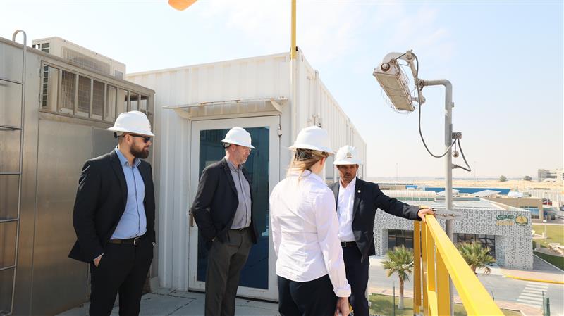 Four people in hard hats at an industrial site, standing on a platform near equipment and a building.